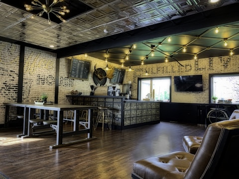 A modern cafe interior with industrial-style decor. The space features wooden flooring and a high ceiling with geometric lighting fixtures. A long counter with a series of small drawers runs alongside one wall, topped with a menu board and coffee-making equipment. Seating includes leather chairs and a long communal table with benches. Exposed brick walls add texture, while large windows provide natural light.