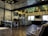 A modern cafe interior with industrial-style decor. The space features wooden flooring and a high ceiling with geometric lighting fixtures. A long counter with a series of small drawers runs alongside one wall, topped with a menu board and coffee-making equipment. Seating includes leather chairs and a long communal table with benches. Exposed brick walls add texture, while large windows provide natural light.
