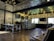 A modern cafe interior with industrial-style decor. The space features wooden flooring and a high ceiling with geometric lighting fixtures. A long counter with a series of small drawers runs alongside one wall, topped with a menu board and coffee-making equipment. Seating includes leather chairs and a long communal table with benches. Exposed brick walls add texture, while large windows provide natural light.