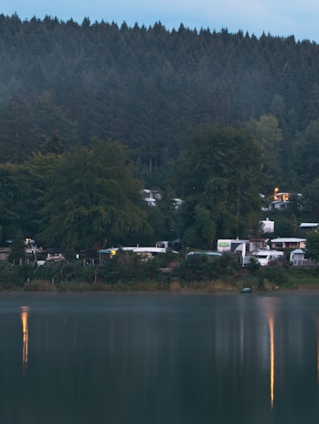 A serene lakeside scene with dense evergreen forest in the background and a calm body of water in the foreground. Along the shore, several small cabins or RVs are nestled among the trees with soft lights visible from the windows. The surface of the water reflects a few lights from the dwellings, enhancing the tranquil atmosphere.