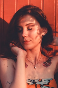 A woman with a serene expression closes her eyes, framed by shadows and soft sunlight. She has floral tattoos on her shoulders and wears a crescent moon necklace. Her dark hair contrasts with the reddish-brown background.