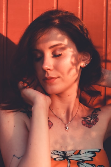 A woman with a serene expression closes her eyes, framed by shadows and soft sunlight. She has floral tattoos on her shoulders and wears a crescent moon necklace. Her dark hair contrasts with the reddish-brown background.