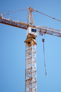 A tall construction crane with a lattice structure extends high into the clear blue sky. The crane is equipped with a pulley system, and a cable is hanging down from the end.