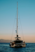 A catamaran with a tall mast floats on a calm sea during sunset. The sky transitions from blue to a soft orange near the horizon. Mountains are visible in the background, adding to the serene setting.