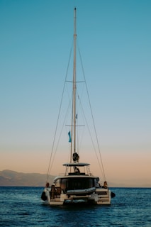 A catamaran with a tall mast floats on a calm sea during sunset. The sky transitions from blue to a soft orange near the horizon. Mountains are visible in the background, adding to the serene setting.