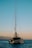 A catamaran with a tall mast floats on a calm sea during sunset. The sky transitions from blue to a soft orange near the horizon. Mountains are visible in the background, adding to the serene setting.