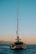 A catamaran with a tall mast floats on a calm sea during sunset. The sky transitions from blue to a soft orange near the horizon. Mountains are visible in the background, adding to the serene setting.