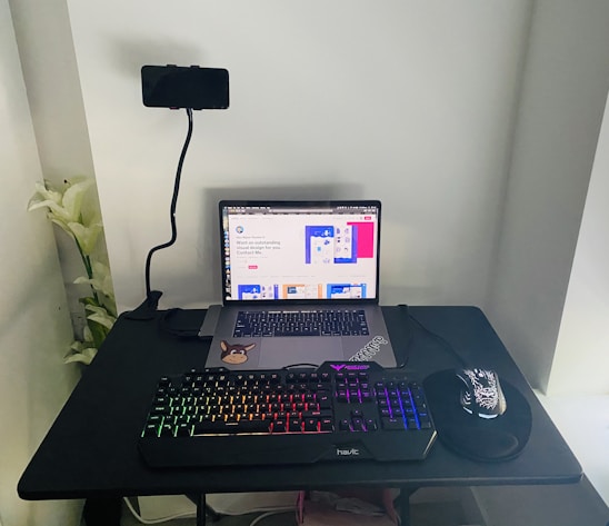 A home office setup with a laptop on a black desk, displaying a webpage. A mechanical keyboard with rainbow backlighting, a mouse with a floral pattern on a mousepad, and a phone mounted on a flexible stand are also present. White flowers are visible on the left side.