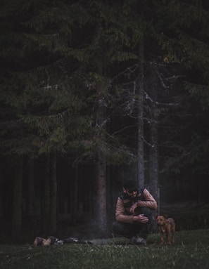A person crouches near a small dog in a dark, dense forest. There is a faint smoke rising from a campfire on the ground. The person is wearing a jacket and appears to be interacting with the dog. The tall, thick trees create a secluded and mysterious atmosphere.