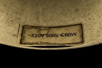 A close-up view of a label with electrical specifications stating approximately 220V, 50Hz, and maximum 60W. The label is affixed to a slightly textured, metallic surface and is set against a dark background.