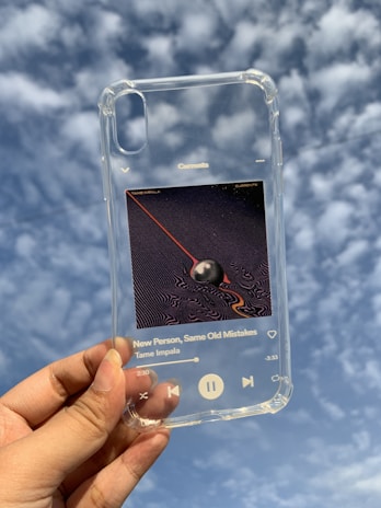 A transparent smartphone case is being held up against a cloudy sky. The case features an album cover design, with a black sphere and red lines against a patterned background. Song information displayed includes the title 'New Person, Same Old Mistakes' by Tame Impala.