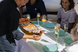 A person is serving a platter shaped like a boat filled with various sushi rolls to a group of people seated at a table. The table is set with blue plates and glasses filled with beverages, including champagne and orange juice. The group includes people of different ages dressed in casual clothing.