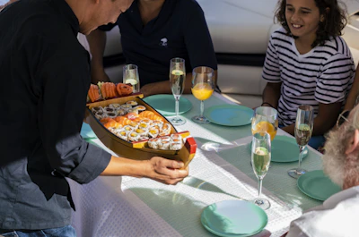 A person is serving a platter shaped like a boat filled with various sushi rolls to a group of people seated at a table. The table is set with blue plates and glasses filled with beverages, including champagne and orange juice. The group includes people of different ages dressed in casual clothing.