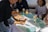 A person is serving a platter shaped like a boat filled with various sushi rolls to a group of people seated at a table. The table is set with blue plates and glasses filled with beverages, including champagne and orange juice. The group includes people of different ages dressed in casual clothing.