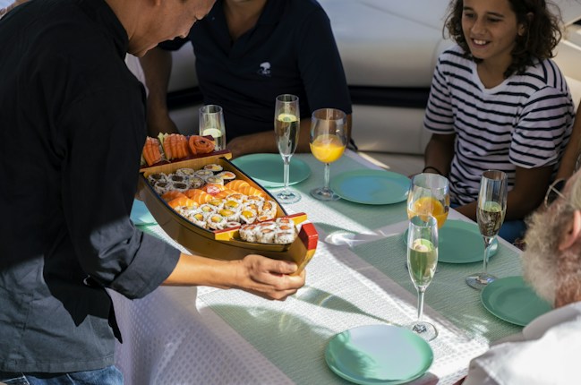 A person is serving a platter shaped like a boat filled with various sushi rolls to a group of people seated at a table. The table is set with blue plates and glasses filled with beverages, including champagne and orange juice. The group includes people of different ages dressed in casual clothing.