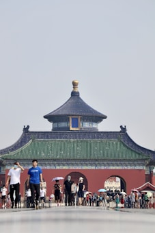 A traditional Chinese temple with an ornate roof and a central tower stands prominently in the background. The structure features intricate designs with green, red, and blue colors. In the foreground, numerous people are walking, some carrying umbrellas for shade. The scene appears busy, with people engaging in leisure or tourism activities.