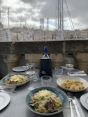 A table set with three plates of pasta dishes garnished with cheese in a restaurant overlooking a marina with sailboats. A bottle of wine and two wine glasses are placed on the table, alongside napkins and cutlery. The background features a cityscape of buildings under a cloudy sky.