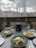 A table set with three plates of pasta dishes garnished with cheese in a restaurant overlooking a marina with sailboats. A bottle of wine and two wine glasses are placed on the table, alongside napkins and cutlery. The background features a cityscape of buildings under a cloudy sky.