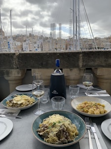 A table set with three plates of pasta dishes garnished with cheese in a restaurant overlooking a marina with sailboats. A bottle of wine and two wine glasses are placed on the table, alongside napkins and cutlery. The background features a cityscape of buildings under a cloudy sky.