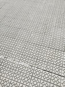 Interlocking stone tiles with a floral pattern cover a large surface, creating a geometric and repetitive design. The light gray stones are arranged in a grid, each featuring a flower motif etched into the surface.