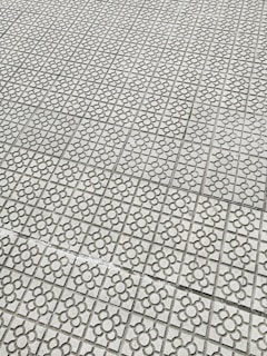 Interlocking stone tiles with a floral pattern cover a large surface, creating a geometric and repetitive design. The light gray stones are arranged in a grid, each featuring a flower motif etched into the surface.