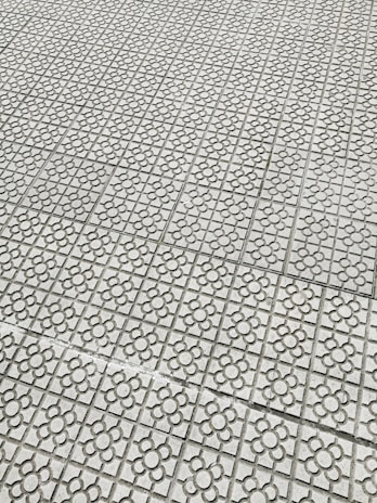 Interlocking stone tiles with a floral pattern cover a large surface, creating a geometric and repetitive design. The light gray stones are arranged in a grid, each featuring a flower motif etched into the surface.