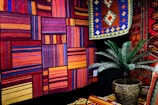 A vibrant display of colorful textiles featuring abstract, geometric patterns. The woven rugs and tapestries hang on the walls and are arranged in bold combinations of reds, oranges, purples, and blues. A large decorative plant in a wicker pot is placed in the foreground, adding a touch of greenery to the scene.