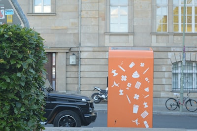 An orange recycling or waste bin with white icons depicting various types of waste, such as cigarette butts, drink cups, and banana peels, is placed in an urban setting. In the background, a parked black car, a bicycle, and a building with large windows are visible. Green foliage partially obscures the scene on the left side.
