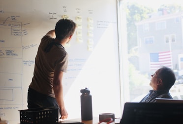 Two people are engaged in a brainstorming session in front of a whiteboard filled with diagrams and sticky notes. One person is writing on the board, while the other is seated and observing. Sunlight streams through a window, casting light onto the scene.