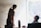 Two people are engaged in a brainstorming session in front of a whiteboard filled with diagrams and sticky notes. One person is writing on the board, while the other is seated and observing. Sunlight streams through a window, casting light onto the scene.