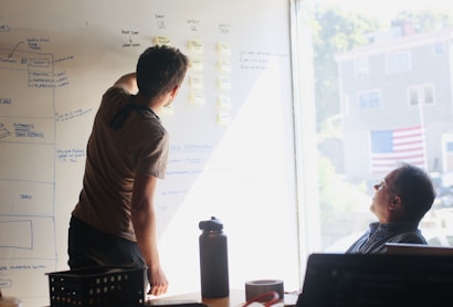 Two people are engaged in a brainstorming session in front of a whiteboard filled with diagrams and sticky notes. One person is writing on the board, while the other is seated and observing. Sunlight streams through a window, casting light onto the scene.