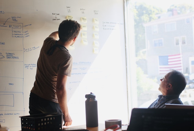 Two people are engaged in a brainstorming session in front of a whiteboard filled with diagrams and sticky notes. One person is writing on the board, while the other is seated and observing. Sunlight streams through a window, casting light onto the scene.
