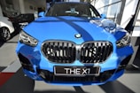 A bright blue BMW X1 parked indoors, showcasing its prominent grille and sleek design in a well-lit showroom with other cars visible in the background.
