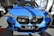 A bright blue BMW X1 parked indoors, showcasing its prominent grille and sleek design in a well-lit showroom with other cars visible in the background.