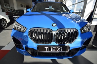 A bright blue BMW X1 parked indoors, showcasing its prominent grille and sleek design in a well-lit showroom with other cars visible in the background.