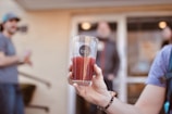 A hand holds a pint glass filled with a reddish beverage, with the logo 'tool' visible on the glass. The background is softly blurred, showing two people in casual attire standing near a building entrance.