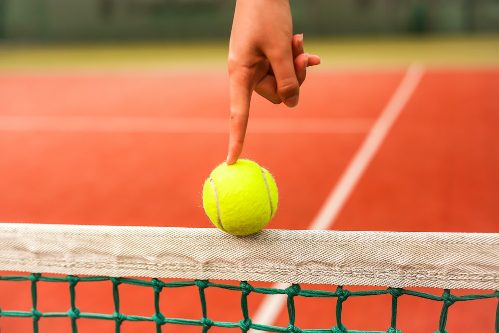 Persona sosteniendo una pelota de tenis amarilla en la red roja y blanca