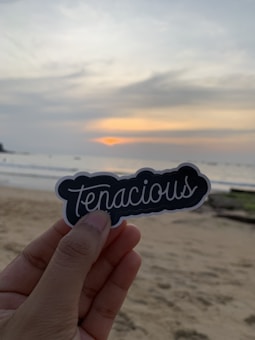 A hand holding a sticker with the word 'tenacious' written on it against the backdrop of a sandy beach and a serene sunset over the ocean, with soft clouds in the sky.