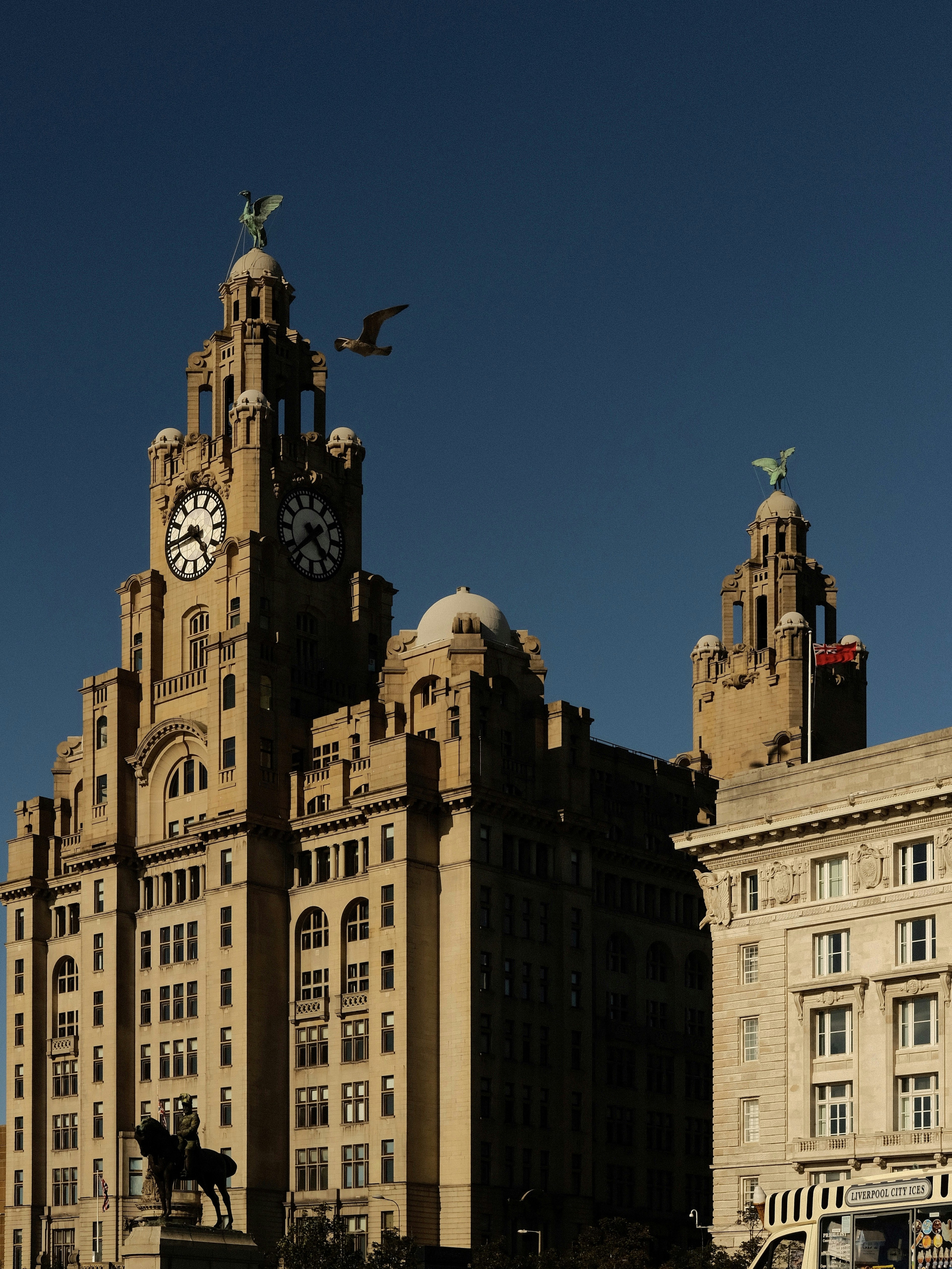 Liverpool's Royal Liver Building rises with twin clock towers against a deep blue sky, with seagulls hovering above and a horse statue at street level.