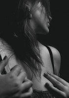 A black and white portrait of a woman captured from a side angle, highlighting her shoulder and part of her face. Her hair falls loosely over her shoulder, and her hand is delicately placed on her chest. The image focuses on the textures of her skin and hair, creating a dramatic and intimate mood.