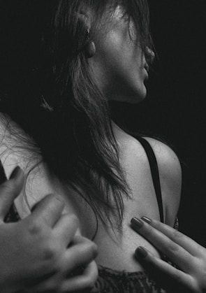 A black and white portrait of a woman captured from a side angle, highlighting her shoulder and part of her face. Her hair falls loosely over her shoulder, and her hand is delicately placed on her chest. The image focuses on the textures of her skin and hair, creating a dramatic and intimate mood.