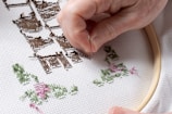 A hand is delicately working on a piece of cross-stitch embroidery using a needle and colorful threads. The fabric is stretched over an embroidery hoop, featuring patterns in brown, green, and pink thread against a white background.