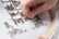 A hand is delicately working on a piece of cross-stitch embroidery using a needle and colorful threads. The fabric is stretched over an embroidery hoop, featuring patterns in brown, green, and pink thread against a white background.