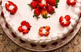 A beautifully decorated cake topped with fresh strawberries arranged in symmetrical patterns. The cake has a layer of white frosting, with piped decorations around the edges. The surface on which the cake sits appears to be a speckled countertop.