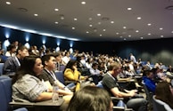 A large group of people seated in an auditorium, attentively listening. The room is dimly lit with spotlights on the dark blue walls, creating a focused atmosphere. Most attendees are casually dressed and some are taking notes or engaging with devices.