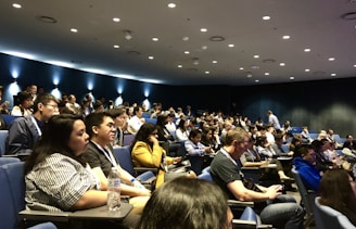 A large group of people seated in an auditorium, attentively listening. The room is dimly lit with spotlights on the dark blue walls, creating a focused atmosphere. Most attendees are casually dressed and some are taking notes or engaging with devices.