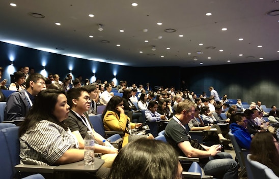 A large group of people seated in an auditorium, attentively listening. The room is dimly lit with spotlights on the dark blue walls, creating a focused atmosphere. Most attendees are casually dressed and some are taking notes or engaging with devices.