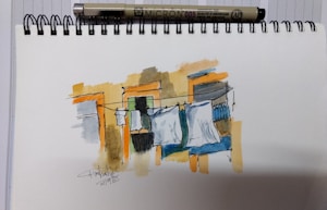 A sketch displays a scene of clothes hanging on a line against the backdrop of a building. The artwork, done in ink and watercolor, incorporates minimalistic strokes with shades of orange, beige, and blue. A Micron pen lies atop the spiral-bound sketchpad which features a signature and date.