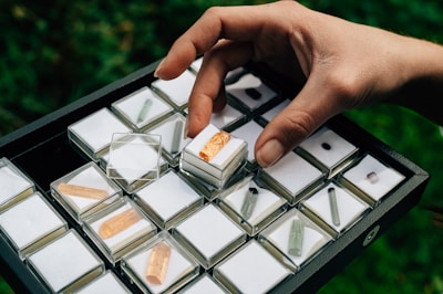 A hand is holding a box with a small orange crystal. The box is placed within a larger tray that is filled with similar small boxes, each containing different colored crystals with transparent lids. The background is blurred and green, suggesting an outdoor setting.