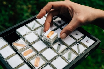A hand is holding a box with a small orange crystal. The box is placed within a larger tray that is filled with similar small boxes, each containing different colored crystals with transparent lids. The background is blurred and green, suggesting an outdoor setting.